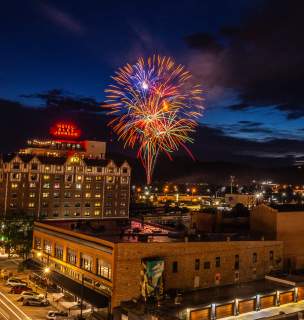 Fireworks burst in the night sky above downtown Rapid City, with the illuminated Hotel Alex Johnson and city lights glowing beneath dark clouds at dusk.