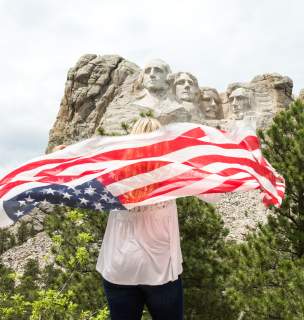 A woman holding an American flag behind her faces away from the camera toward Mount Rushmore, visible in the upper half of the photo under a mostly cloudy sky.