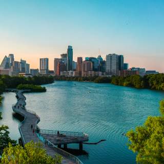 People walking along suspended board walk that follows the shape of the Colorado River, leading to Downtown Austin skyline.