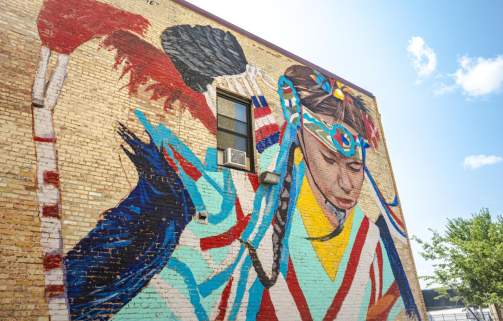Coming Home mural of Native American women on the side of a tan brick building.