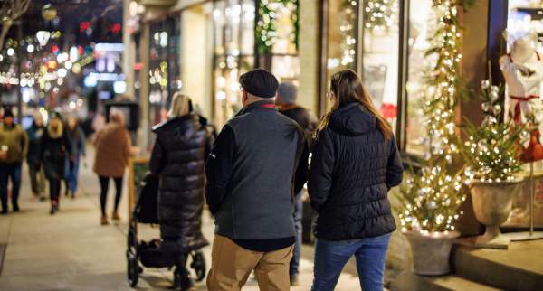 People walk along a city sidewalk decorated with holiday lights and storefront displays at night
