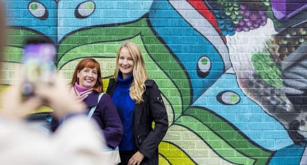 two women posing for a cell phone photo in front of a colorful mural of a bird