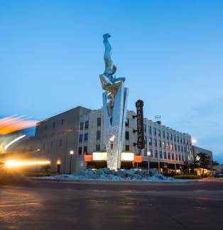 steel sculpture "muskegon rising" sits in roundabout in front of historic Frauenthal theater at sunset. blurred car headlights illuminate the left side of the photo