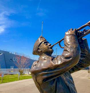 A bronze statue of a person playing a trumpet, with the USS LST 393 Museum in the background under a clear blue sky.