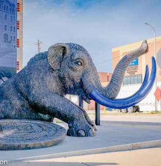A large sculpture of a mastodon with blue tusks is placed outside the Lakeshore Museum Center. The creature is crouched on a circular base, and a "Visit Muskegon" logo is seen in the corner. The background includes a brick building and a delivery truck.