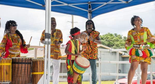 Five people dressed in brightly colored clothes, honoring their ethnicity, playing various percussion instruments on an outdoor stage, under a blue canopy.