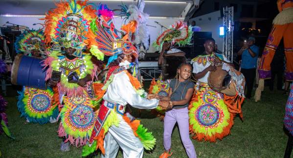 A group dressed in brightly-colored traditional Caribbean Festival attire dancing with a young girl and playing drums in front of an outdoor stage in Pompano Beach