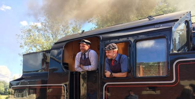 A driver and his crew smiling as they stand in the engine of the Bluebell Railway steam train