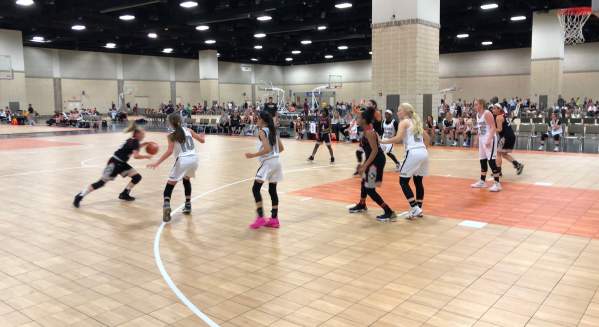 A high-energy basketball game unfolds in a large indoor arena, with players in white, black, and pink jerseys competing for the ball on a wooden court.