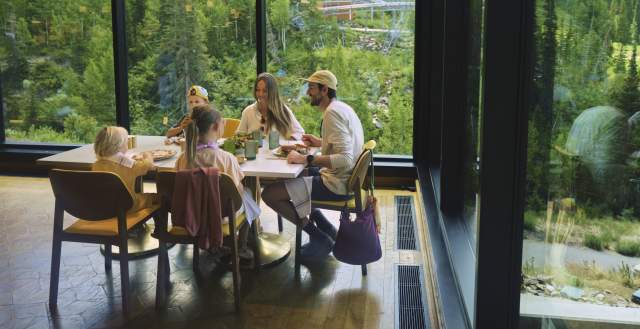 Family dining at an indoor table with very large glass windows all around looking out at the side of a mountain covered in green pine trees