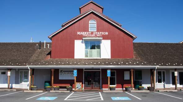 Market Station red barn-style building with a sloped roof, surrounded by parking spaces and planters. Clear blue sky above.