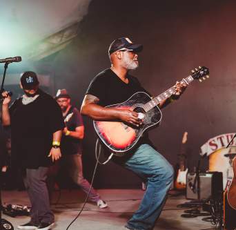 Musician Ray Prim plays an acoustic sunburst guitar on stage at Stubb's in Austin Texas. Two other male musicians stand behind him