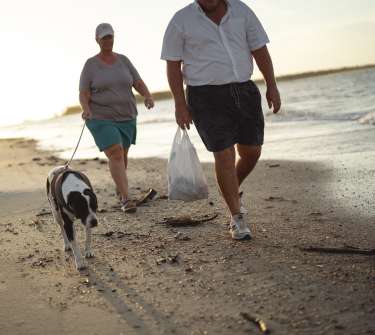 a man and woman walking a dog on the beach carrying a trash bag