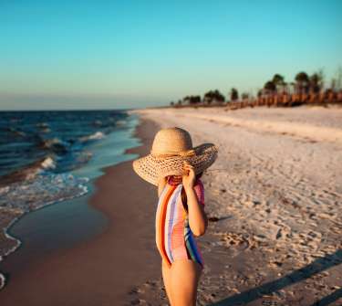 a little girl wearing a striped bathing suit and straw hat on a beach with a moody feel and a blue sky