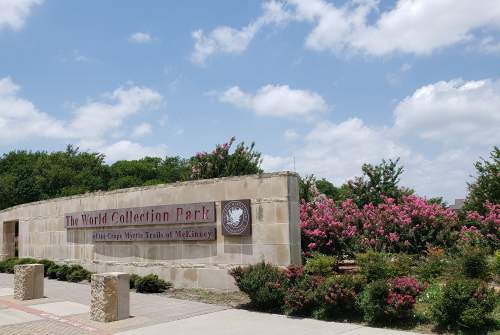Sign for The World Collection Park, surrounded by blooming pink crape myrtle trees and greenery under a partly cloudy sky.
