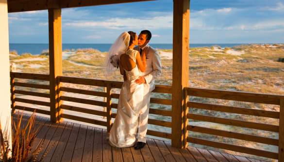 A couple on their wedding day kiss on a porch overlooking the beach in the Outer Banks