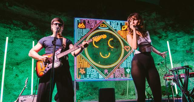 Man playing guitar and woman singing into a microphone on a stage backlit by a neon smiley face.
