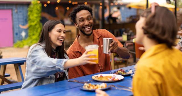 Two women and a man cheering drinks over plates of tacos.