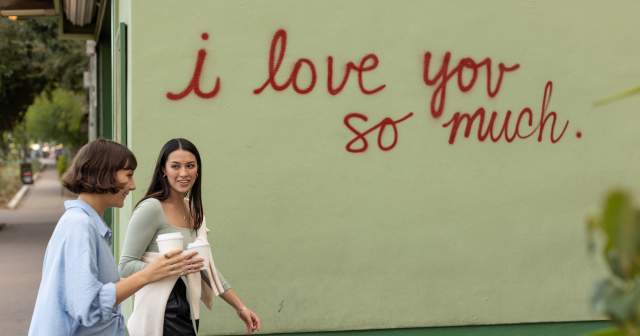 Two women walking in front of the I Love You So Much mural with coffee in hand.
