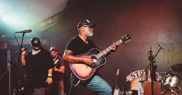 Musician Ray Prim plays an acoustic sunburst guitar on stage at Stubb's in Austin Texas. Two other male musicians stand behind him