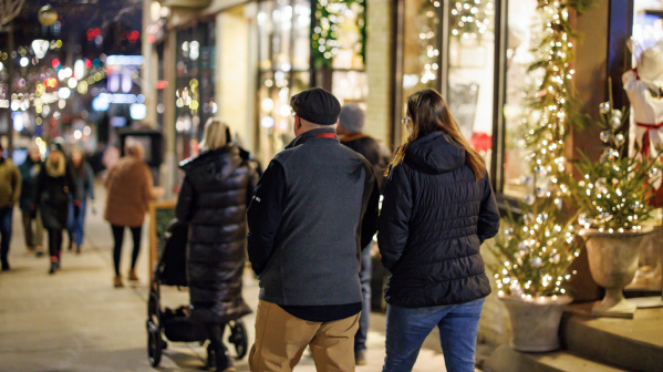 People walk along a city sidewalk decorated with holiday lights and storefront displays at night