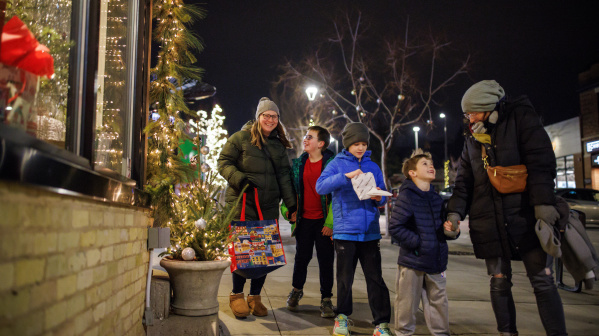 A group of two adults and three children walk together on a decorated city sidewalk at night during the holiday season. They are bundled in winter coats, hats, and gloves. Festive lights and small decorated trees line the shop windows on the left. One adult carries a colorful tote bag, and one child holds a snack. Everyone appears cheerful and engaged with one another as they enjoy the winter evening.”