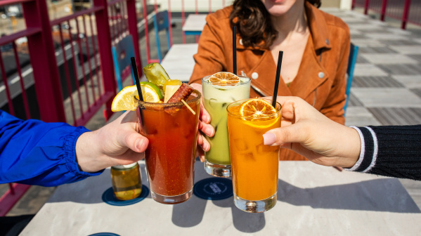 Three people clinking colorful cocktails on a rooftop patio, featuring a Bloody Mary, a green drink, and an orange drink, with a bright and casual vibe.