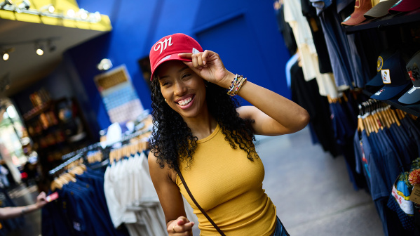 Smiling shopper trying on a red Miller-branded baseball cap inside the Miller Brewery gift shop, surrounded by racks of apparel and branded merchandise.