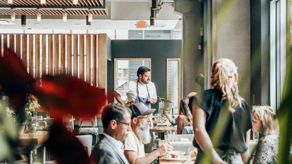 Diners enjoy a meal inside a bright, modern Milwaukee restaurant with large windows, warm lighting, and a relaxed, welcoming atmosphere as a chef engages with guests.