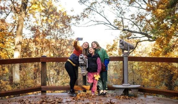 Family smiles and poses for a selfie outdoors during a fall day.