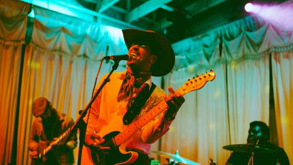 Musician in a cowboy hat singing into a mic while playing guitar with colorful lights surrounding him.