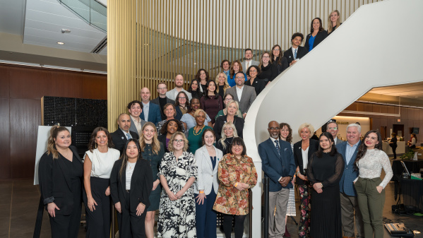 Large group of professionals posing on a curved staircase inside a modern building with wood slat walls and glass railings.