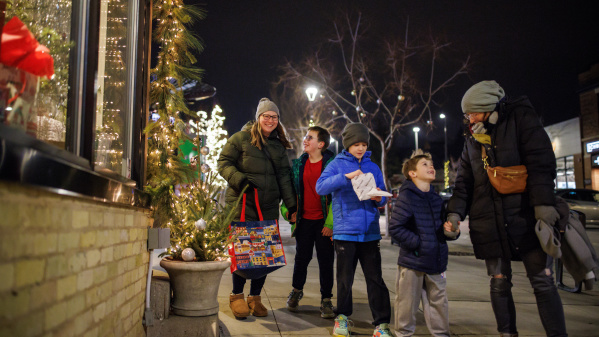 A group of two adults and three children walk together on a decorated city sidewalk at night during the holiday season. They are bundled in winter coats, hats, and gloves. Festive lights and small decorated trees line the shop windows on the left. One adult carries a colorful tote bag, and one child holds a snack. Everyone appears cheerful and engaged with one another as they enjoy the winter evening.”