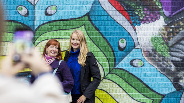 two women posing for a cell phone photo in front of a colorful mural of a bird