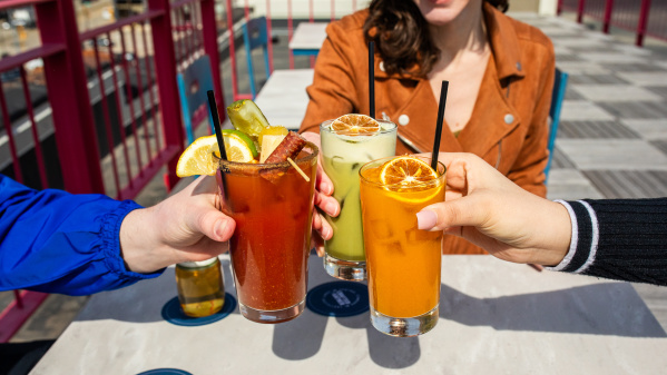 Three people clinking colorful cocktails on a rooftop patio, featuring a Bloody Mary, a green drink, and an orange drink, with a bright and casual vibe.