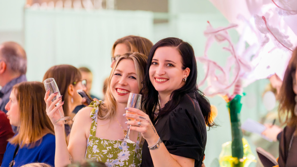 Two women smiling and posing together at a crowded indoor event, each holding a champagne flute, with floral decorations and other guests in the background.