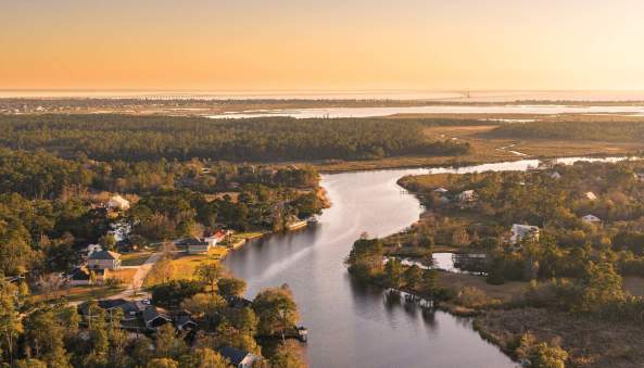 Aerial sunset view of a winding Slidell waterway bordered by homes, marshes and trees, with Lake Pontchartrain stretching across the horizon on the Fall/Winter 2026–27 cover of The Northshore.