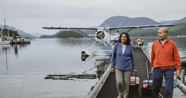 Two people wheel their suitcases along the dock after disembarking from a floatplane.