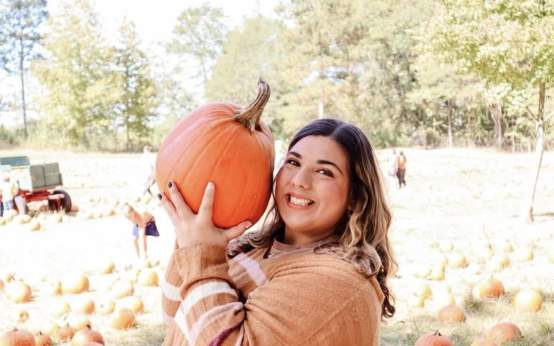 A smiling girl holds a pumpkin in front of a pumpkin patch.