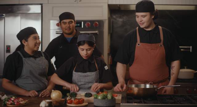 Four student chefs standing in a designed commercial kitchen, three of the students watching one of them chopping a vegetable.