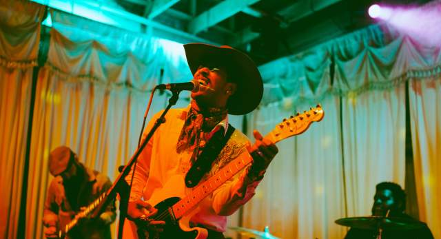 Musician in a cowboy hat singing into a mic while playing guitar with colorful lights surrounding him.