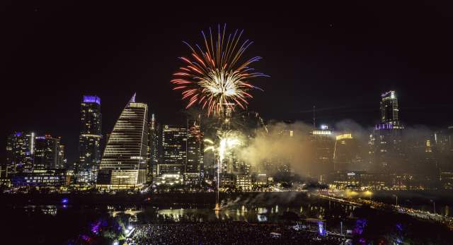 Red fireworks explode over downtown Austin skyline and Lady Bird Lake at night. Aerial view shows a music stage and crowd gathered below at Auditorium shores.