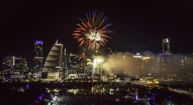 Red fireworks explodes over downtown Austin skyline and Lady Bird Lake at night. Aerial view shows a music stage and crowd gathered below at Auditorium shores.