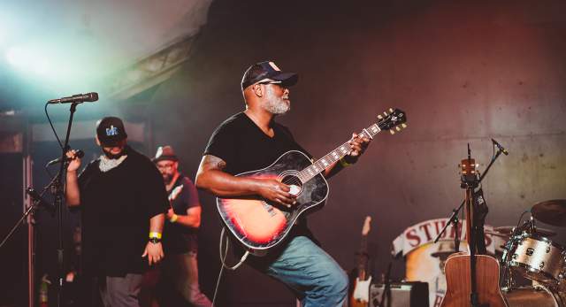 Musician Ray Prim plays an acoustic sunburst guitar on stage at Stubb's in Austin Texas. Two other male musicians stand behind him