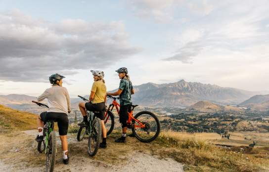 Three mountain bikers at the top of a mountain overlooking a valley with the Remarkables mountains in the background