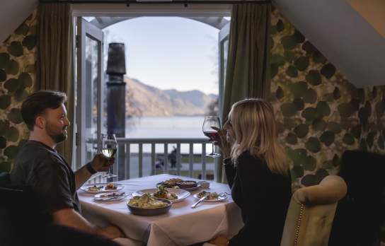 A cosy dinner setting with a couple enjoying wine and a delicious meal, overlooking stunning lake and mountain views through open balcony doors.