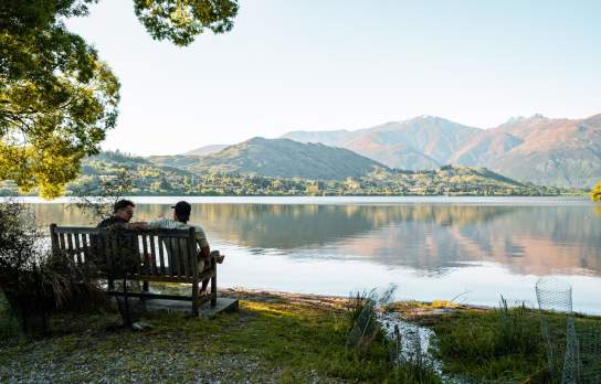 Two people sitting on a bench looking our over a lake with mountains in the background