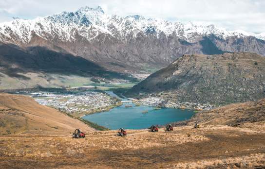 A scenic view of Queenstown, New Zealand, captured from a high vantage point. The image shows snow-capped mountains in the background, a town nestled by a serene lake in the valley, and a group of quad bikes traversing a grassy hill in the foreground.