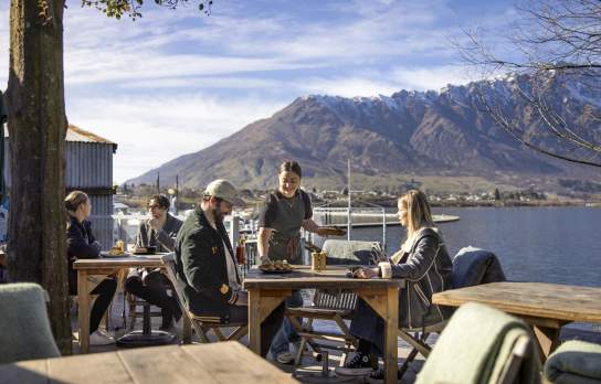 People dining outside next to the lake with the Remarkables mountain range in the background at The Boat Shed