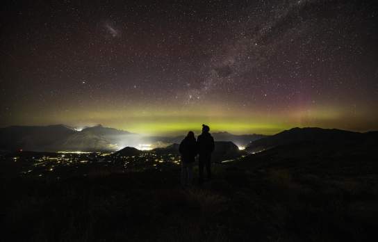 People standing on a mountain top with a starry night sky and aroura before them